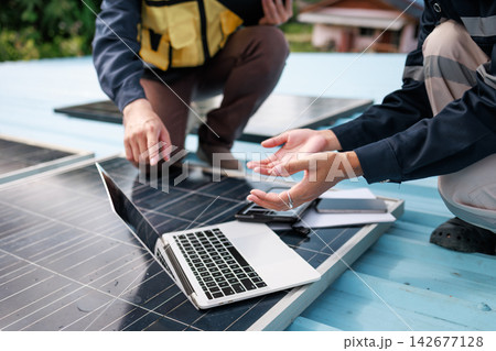 Two technicians inspect solar panels on a rooftop beside a laptop, planning renewable energy installation and maintenance outdoors. 142677128