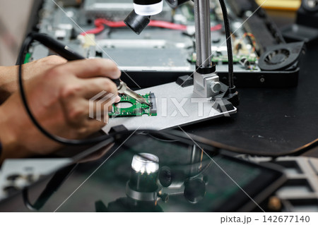 Technician repairs a green circuit board with a soldering iron under a magnifying lamp at an electronics workbench. Technician repairs a green circuit board with a soldering iron under a magnifying lamp at an electronics workbench. 142677140