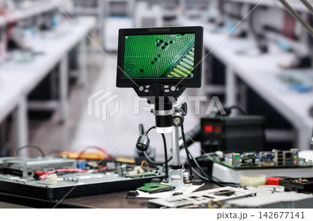 Digital microscope on a workbench displays a close view of a green circuit board in a busy electronics repair workshop. Digital microscope on a workbench displays a close view of a green circuit board in a busy electronics repair workshop. 142677141