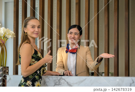 Happy female guest giving thumbs up at hotel reception front desk while receptionist provides assistance Happy female guest giving thumbs up at hotel reception front desk while receptionist provides assistance 142678319