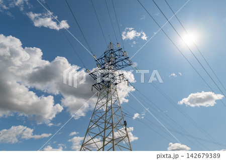 Electric power lines stretch high against a bright blue sky filled with fluffy white clouds on a sunny day Electric power lines stretch high against a bright blue sky filled with fluffy white clouds on a sunny day 142679389