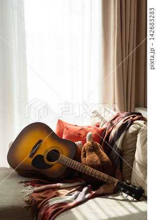 Acoustic guitar resting on a sofa with cushions and a plaid blanket near a window with sheer curtains creating soft lighting. Acoustic guitar resting on a sofa with cushions and a plaid blanket near a window with sheer curtains creating soft lighting. 142683283