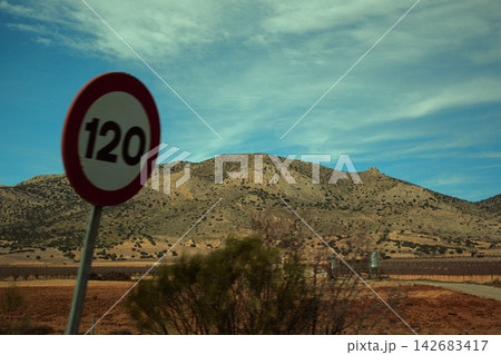 Speed limit sign with number 120 on rural road passing dry mountainous landscape under blue sky 142683417