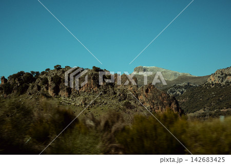 Expansive rugged mountains featuring rocky outcrops, green scrub, and sparse trees beneath a vast, serene blue sky Expansive rugged mountains featuring rocky outcrops, green scrub, and sparse trees beneath a vast, serene blue sky 142683425