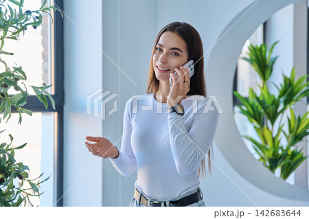 Smiling young woman talking on smartphone looking at camera in modern interior Smiling young woman talking on smartphone looking at camera in modern interior 142683644