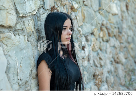 Young woman with long black hair leaning against stone wall 142683706