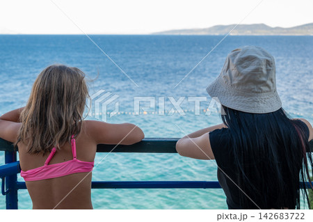 Two women relaxing at seaside railing while overlooking calm blue water Two women relaxing at seaside railing while overlooking calm blue water 142683722