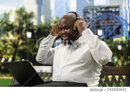 Professional man adjusting headphones on video call remote while seated with laptop on park bench, preparing to connect or listen during flexible work outdoors. 142685641
