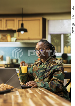 African american woman soldier off duty in the kitchen using laptop during morning routine. Enjoying coffee cup and pastry for a domestic life in a calm quiet environment at home. African american woman soldier off duty in the kitchen using laptop during morning routine. Enjoying coffee cup and pastry for a domestic life in a calm quiet environment at home. 142685670