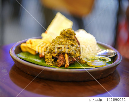 Traditional Indonesian duck dish (bebek sambal ijo) topped with rich green chili sambal, served alongside steamed white rice, fried tofu, tempeh, and fresh cucumber garnish on a traditional banana lea Traditional Indonesian duck dish (bebek sambal ijo) topped with rich green chili sambal, served alongside steamed white rice, fried tofu, tempeh, and fresh cucumber garnish on a traditional banana lea 142690032