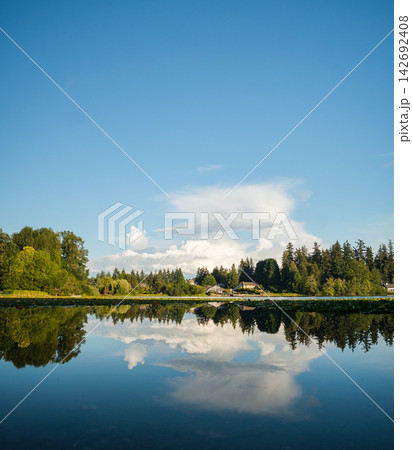 Perfect mirror reflection of trees and clouds on a calm lake surface under a blue sky. Perfect mirror reflection of trees and clouds on a calm lake surface under a blue sky. 142692408