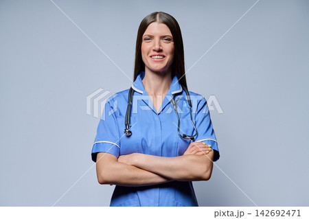 Confident female nurse in blue uniform with stethoscope standing with arms crossed against grey background Confident female nurse in blue uniform with stethoscope standing with arms crossed against grey background 142692471