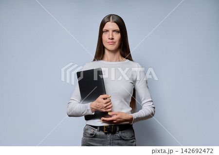 Portrait of serious young woman holding closed laptop computer on grey background Portrait of serious young woman holding closed laptop computer on grey background 142692472