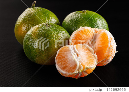 Fresh green tangerines with one peeled open showing juicy orange segments against black textured background 142692666