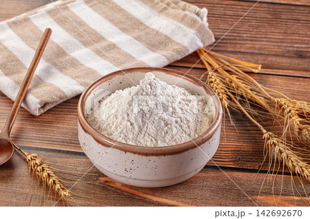 Bowl of white flour on rustic wooden table with wheat stalks, striped cloth, and wooden spoon, warm natural lighting, shallow depth of field 142692670