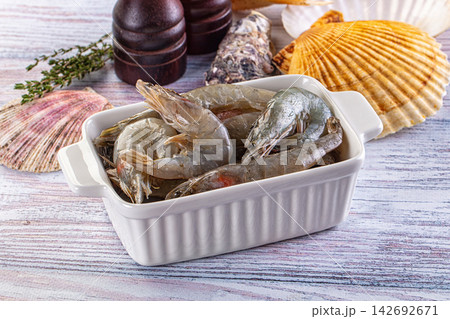 Raw fresh shrimp in white ceramic dish, green thyme sprig, variety of seashells, wooden grinders on rustic light wood background, seafood preparation 142692671