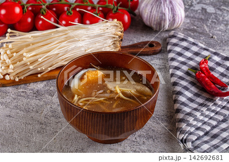 Bowl of miso ramen with soft boiled egg, enoki mushrooms and chicken in rich broth, surrounded by cherry tomatoes, garlic, red chili peppers and checkered napkin on gray stone surface 142692681
