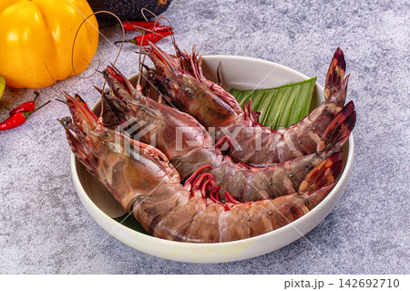 Fresh raw tiger prawns in white bowl with banana leaf, yellow bell pepper and red chili peppers on textured gray surface 142692710