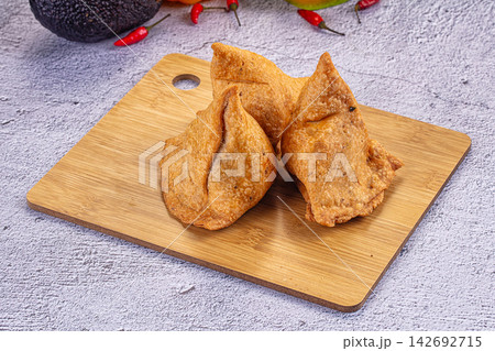 Golden brown samosas on wooden cutting board with red chili peppers and avocado in background, rustic food presentation Golden brown samosas on wooden cutting board with red chili peppers and avocado in background, rustic food presentation 142692715