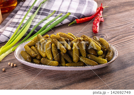 Small pickled gherkins on white serving plate, fresh green onions, red chili peppers, black peppercorns, and checkered kitchen towel on rustic dark wooden table 142692749