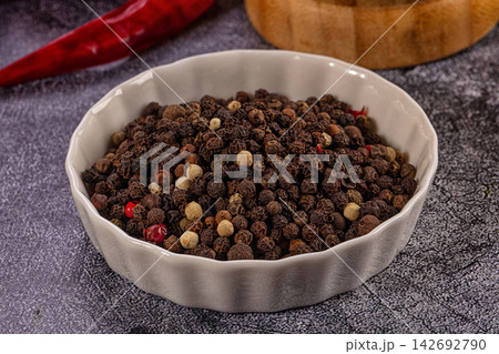 White ceramic bowl filled with mixed peppercorns including black, white, and red, on textured gray surface with red chili and wooden bowl in background White ceramic bowl filled with mixed peppercorns including black, white, and red, on textured gray surface with red chili and wooden bowl in background 142692790