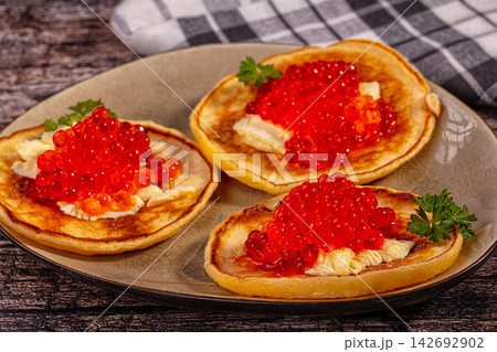 Three golden pancakes topped with butter and bright red caviar, garnished with fresh parsley on rustic ceramic plate 142692902