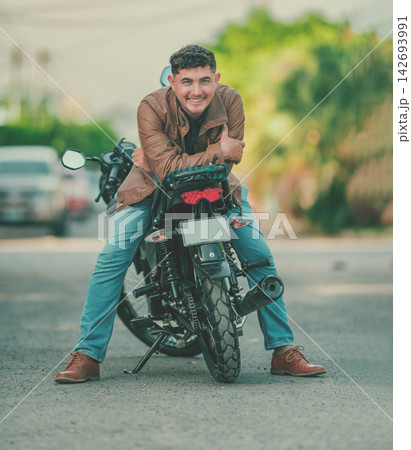 Smiling latin motorcyclist leaning against his motorcycle and looking at the camera 142693991
