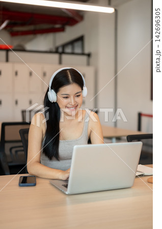 Young Asian woman wearing headphones smiling while typing on laptop at table in a co working cafe. 142696305