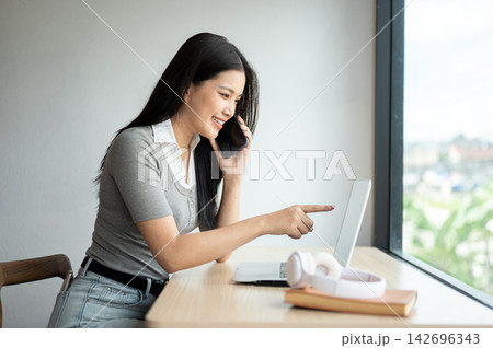 Young Asian woman pointing at a laptop during a phone call while sitting at table in co working cafe Young Asian woman pointing at a laptop during a phone call while sitting at table in co working cafe 142696343