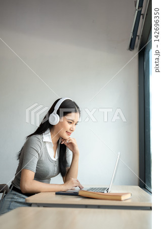 Young Asian woman wearing headphones and using a laptop while sitting at table in a co working cafe. 142696350