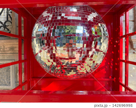 A disco ball is hanging inside a red phone booth located in a busy urban area. 142696998