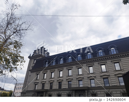 Low angle view of an elegant historic stone building featuring a dark mansard roof with arched dormer windows and a corner spire tower under a cloudy overcast sky. Low angle view of an elegant historic stone building featuring a dark mansard roof with arched dormer windows and a corner spire tower under a cloudy overcast sky. 142698604
