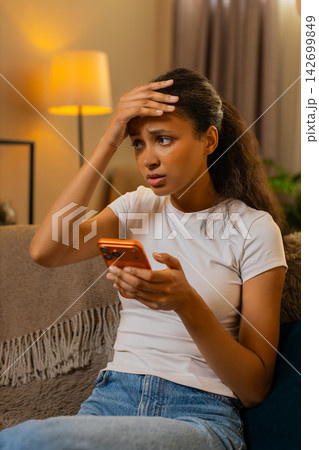 African American young woman on home sofa shows disappointment after reading bad news on smartphone African American young woman on home sofa shows disappointment after reading bad news on smartphone 142699849