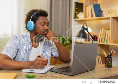African American young man on home table taking online course listening and writing notes for study African American young man on home table taking online course listening and writing notes for study 142699858