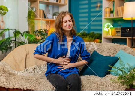 Young woman on home sofa feeling stomach pain and discomfort with painful expression indoors at home Young woman on home sofa feeling stomach pain and discomfort with painful expression indoors at home 142700571