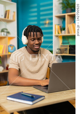 African American young man on home table in headphones working on laptop listening to music for work African American young man on home table in headphones working on laptop listening to music for work 142700920