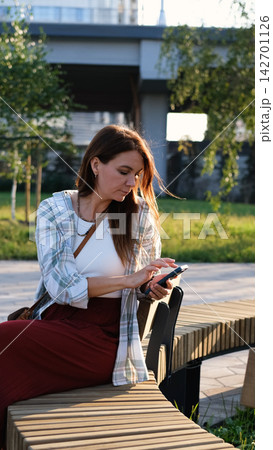Woman Relaxing on a Wooden Park Bench with Smartphone in Warm Golden Evening Sunlight Woman Relaxing on a Wooden Park Bench with Smartphone in Warm Golden Evening Sunlight 142701126