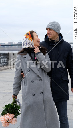 Two adults are kissing on a quiet shore. The woman is holding pale pink flowers and wearing a patterned headscarf. Soft, overcast light, a blurred shoreline, an open sky, and a serene city skyline. Two adults are kissing on a quiet shore. The woman is holding pale pink flowers and wearing a patterned headscarf. Soft, overcast light, a blurred shoreline, an open sky, and a serene city skyline. 142701684