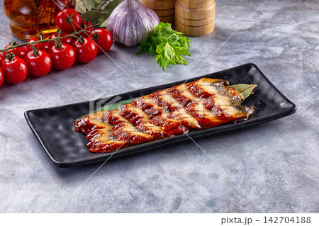 Grilled eel with glossy sauce on black rectangular plate, surrounded by cherry tomatoes, garlic, parsley and wooden spice grinders on gray stone surface Grilled eel with glossy sauce on black rectangular plate, surrounded by cherry tomatoes, garlic, parsley and wooden spice grinders on gray stone surface 142704188