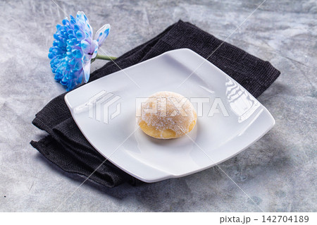 Single mochi dessert dusted with powdered sugar on white plate, blue flower and black napkin on textured gray background Single mochi dessert dusted with powdered sugar on white plate, blue flower and black napkin on textured gray background 142704189