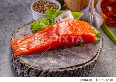 Raw salmon fillet on rustic wooden cutting board with fresh lettuce, sliced onion, garlic, and peppercorns in background 142704256
