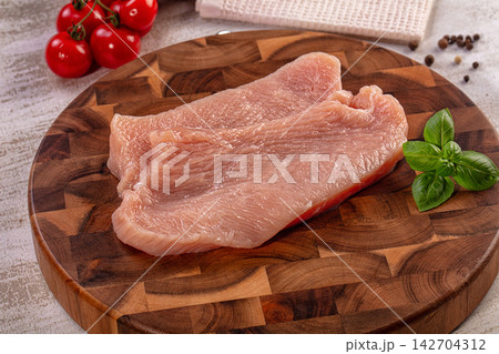Raw turkey breast fillet on wooden cutting board with fresh basil, cherry tomatoes and peppercorns, natural kitchen lighting 142704312