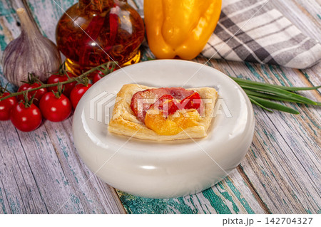 Savory puff pastry tartlet with melted cheese, roasted tomato, and ham on white plate surrounded by fresh garlic, cherry tomatoes, yellow bell pepper, and chives Savory puff pastry tartlet with melted cheese, roasted tomato, and ham on white plate surrounded by fresh garlic, cherry tomatoes, yellow bell pepper, and chives 142704327