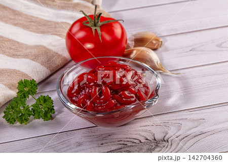 Glass bowl of glossy red ketchup with fresh tomato, garlic cloves, and parsley on light wooden surface beside striped cloth Glass bowl of glossy red ketchup with fresh tomato, garlic cloves, and parsley on light wooden surface beside striped cloth 142704360