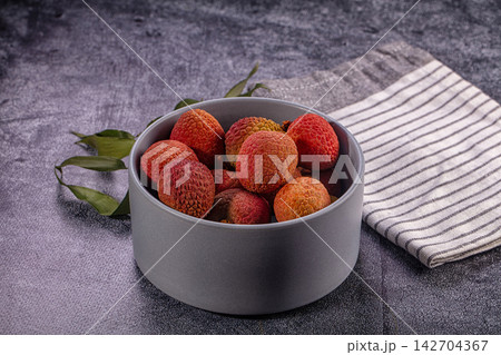 Fresh lychee fruits in gray ceramic bowl with green leaves and striped cloth on dark textured surface 142704367