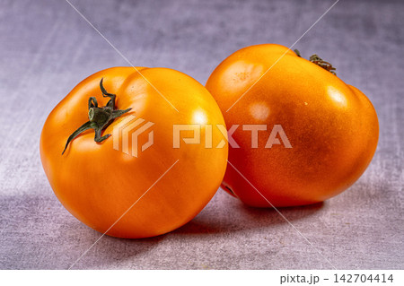 Two ripe orange tomatoes with green stems on textured gray surface, soft studio lighting highlights glossy skin and natural imperfections 142704414