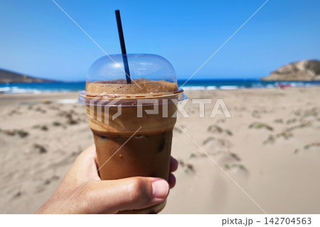Frappe Cold refreshment iced coffee on a sandy beach. Freddo espresso refreshment at summertime. Beach umbrellas in a holiday resort. Frape. 142704563