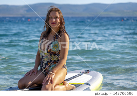 Smiling Woman Sitting on Paddleboard in Calm Water with Mountains 142706596