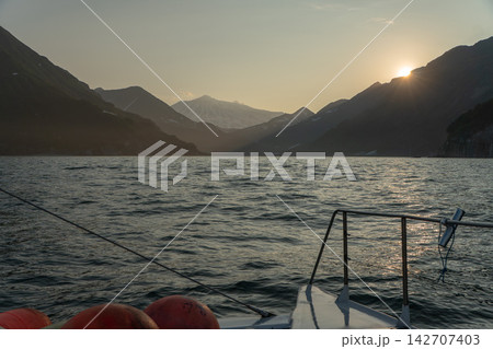 Sun sets behind rugged Kamchatka mountains during boat trip across calm coastal waters. Warm evening light, remote wilderness, marine adventure, scenic journey, tranquility and nature tourism. 142707403