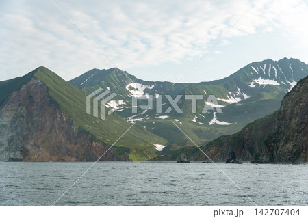 Green mountain ridges and melting snowfields overlook coastal waters in Kamchatka. Remote wilderness, dramatic landscape, scenic marine journey, nature tourism, adventure travel and exploration. Green mountain ridges and melting snowfields overlook coastal waters in Kamchatka. Remote wilderness, dramatic landscape, scenic marine journey, nature tourism, adventure travel and exploration. 142707404